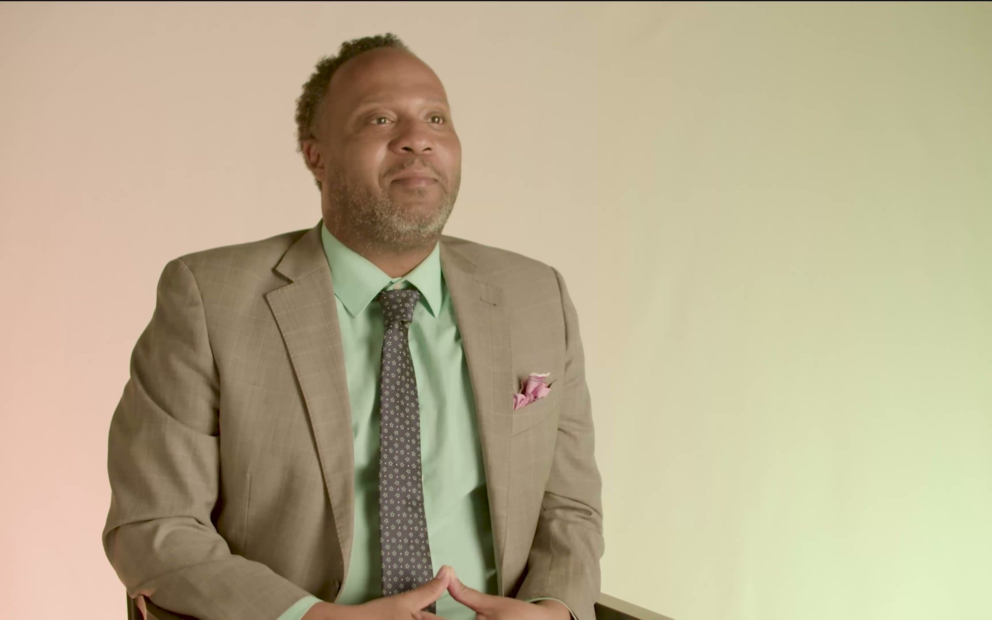 An interview frame: a speaker in a tan suit against a warm cream ground