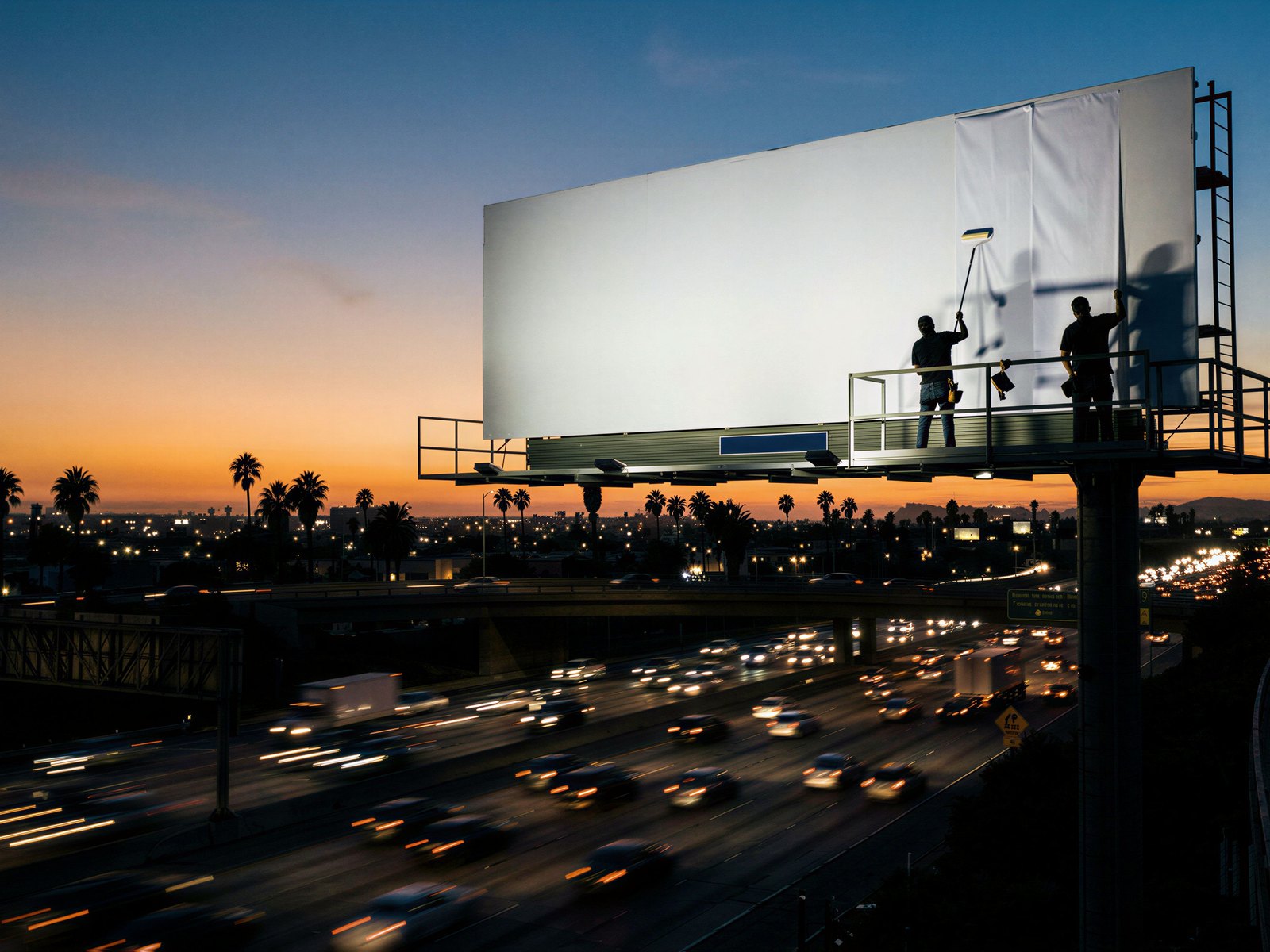 A blank billboard above a Los Angeles freeway at dusk, two workers pasting the poster
