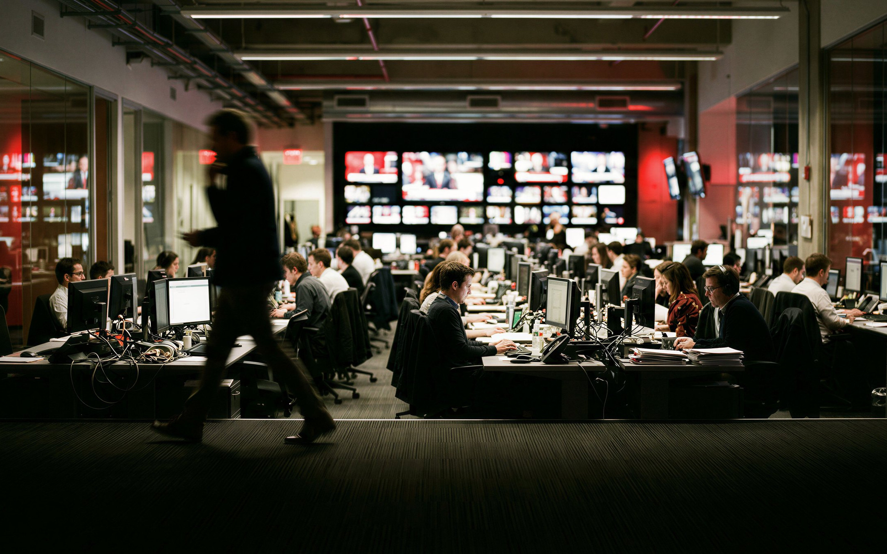 A newsroom at dawn: rows of monitors, a wall of screens, and one person walking past in motion blur